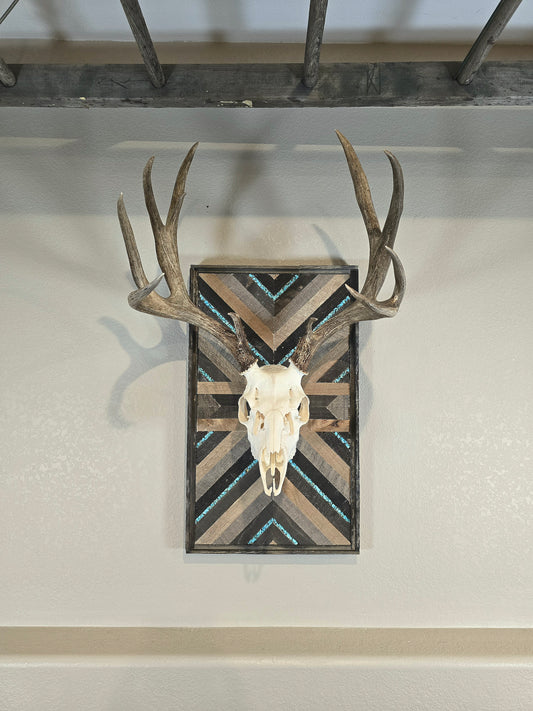 A mounted deer skull on a wall plaque with a geometric pattern, featuring turquoise inlay and wood elements.